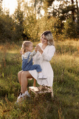 Fototapeta premium Blonde woman and little child standing on sunlit field. Mother in white dress, child in denim overalls. Concept of family, active recreation or children's exploration.