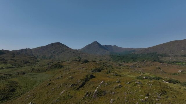 Sugarloaf, Caha Mountains, County Cork, Ireland, September 2024. Drone pushes forward and rises from the craggy landscape filled with rocky crags and grass towards the hills under a clear blue sky.