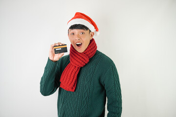 Young asian man wearing santa clause hat, red scarf and green long sleeved sweater is holding dummy credit card over white background.