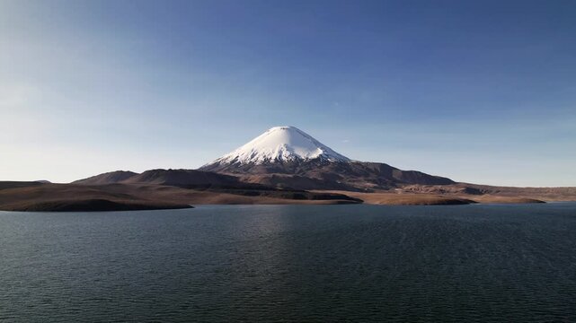 Aerial View Of Vulcan Parinacota In Lauca National Park, Chile, Captured On A Sunny Autumn Day. The Drone Ascends From The Lake, Revealing Snow-Capped Peaks And Sandy Terrain Below.