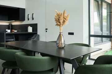 Contemporary dining area showcasing green chairs and a sleek black table with a decorative vase as centerpiece. Modern kitchen elements and large windows complement minimalist design