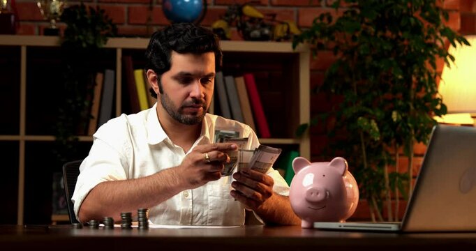 Indian Asian bearded young man sitting at a desk at home, counting Indian currency notes with worried or sad expressions, reflecting financial stress and planning.