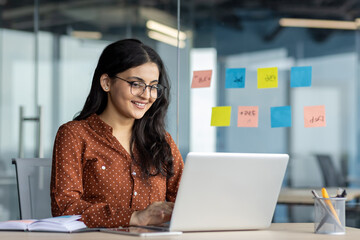 Hispanic woman businesswoman engaged with laptop amid modern office setting. Sticky notes on glass...