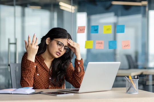 Hispanic businesswoman feeling stressed while working on laptop in office environment, surrounded by notes and papers. Concept of work stress, frustration, and multitasking.