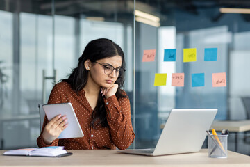 Hispanic woman businesswoman deep in thought in modern office holding tablet. Focused on laptop,...