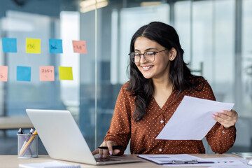 Hispanic woman businesswoman smiles while multitasking with laptop and documents in modern office....