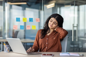 Hispanic woman businesswoman with neck pain sitting in office, laptop open, phone on table. Reflects modern work stress, need for ergonomic solutions, and digital life balance.