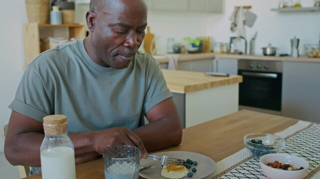 Tilt up shot of middle-aged Black man with diabetes eating low-glycemic healthy breakfast consisting of quark pancakes, fresh blueberries and milk in kitchen at home
