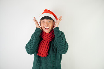 Suprised young asian man wearing santa clause hat, red scarf and green long sleeved sweater win a prize or good news, isolated over white background.