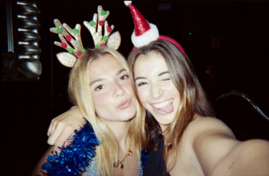 Two young women enjoying a festive Christmas party, wearing holiday accessories and smiling for the camera. The image has a vintage, dusty look with a candid feel.