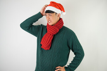 Confused young asian man wearing santa clause hat, red scarf and green long sleeved sweater is scratching his head, isolated over white background.