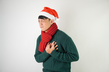 Young asian man wearing santa clause hat, red scarf and green long sleeved sweater  is suffering heart attack and holding his chest, isolated over white background.