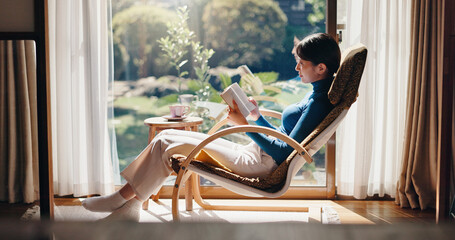 Reading, chill and Asian woman with book, morning and knowledge with literature, relax or weekend. Home, peace and person with novel, rest and comfortable with story, break and cozy on chair or Japan