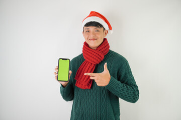 Young asian man wearing santa clause hat, red scarf and green long sleeved sweater is showing smartphone green screen, isolated over white background.