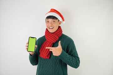 Young asian man wearing santa clause hat, red scarf and green long sleeved sweater is showing smartphone green screen, isolated over white background.
