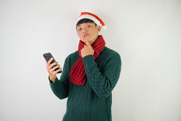 Confused young asian man wearing santa clause hat, red scarf and green long sleeved sweater is using smartphone, isolated over white background.