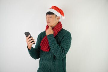 Confused young asian man wearing santa clause hat, red scarf and green long sleeved sweater is using smartphone, isolated over white background.