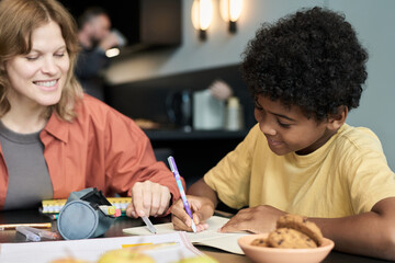 Woman assisting young boy with writing assignments at table with school materials and snacks around. Background shows another person blurred, creating casual atmosphere