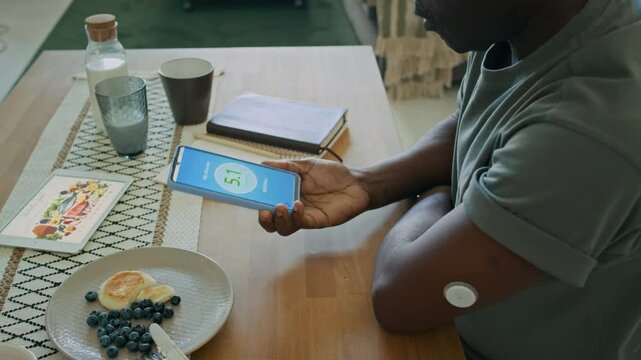 Close up view of Black man with diabetes checking glucose levels with smartphone and CGM sensor, then recording health data in notebook after having breakfast at home
