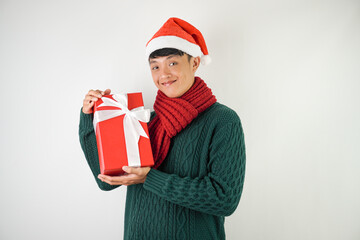 Young asian man wearing santa clause hat, red scarf and green long sleeved sweater with smiling face expression is carrying large gift box with ribbon, isolated over white background.