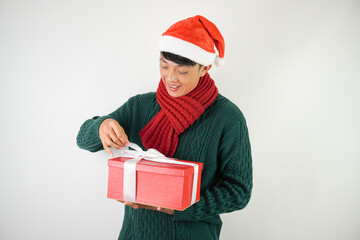 Young asian man wearing santa clause hat, red scarf and green long sleeved sweater with smiling face expression is unboxing large gift box with ribbon, isolated over white background.