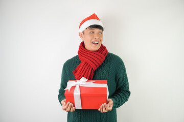 Young asian man wearing santa clause hat, red scarf and green long sleeved sweater with smiling face expression is carrying large gift box with ribbon, isolated over white background.