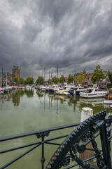 Obraz premium Serene harbor with moored boats under a dramatic cloudy sky. Quaint historic buildings and trees line the waterfront, reflecting in the calm green water, creating peaceful scene. Dordrecht landscape.