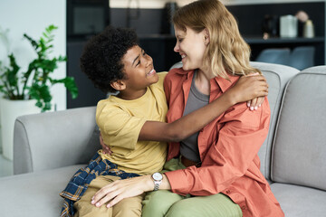 Smiling child wearing yellow shirt embracing caregiver on cozy sofa in modern living room with green plants, creating warm and affectionate atmosphere indoors