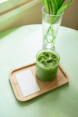 Glass of iced matcha latte with white blank card sits on a wooden tray on a light green table