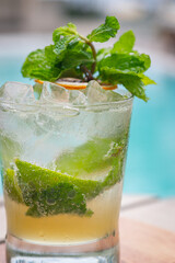 Close up view of Mojito cocktail in glass garnished with a slice of dried lemon and mint leaves on wooden planks in front of a pool