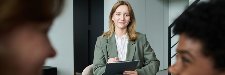 Businesswoman sitting in modern office environment, holding a clipboard, smiling at colleagues. Meeting room features sleek, contemporary design with colleagues blurred in foreground