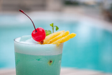 Close up view, Blue hawaii in glass garnished with slice pineapples and cherry and mint leaves on wooden planks in front of a pool