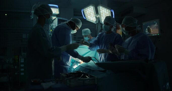 Diverse Surgical Team Working in a Darkened Operating Room Surrounded by Advanced Medical Equipment. Doctors Performing a Complex Surgery with the Help of Laparoscopic Equipment