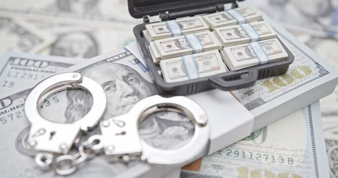 Financial crime, fraud, money laundering, corruption, or law enforcement : Bundles of US dollar 100 bills in a small case, alongside a pair of handcuffs placed over the currency.