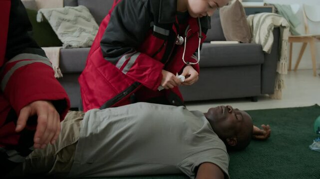 Female paramedic taking tongue depressor from colleague, then examining oral airway of unconscious African American patient lying on floor at home