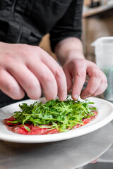 A chef carefully prepares a dish by adding fresh arugula to a plate with thinly sliced meat. The focus is on the meticulous arrangement, showcasing culinary artistry in a kitchen setting.