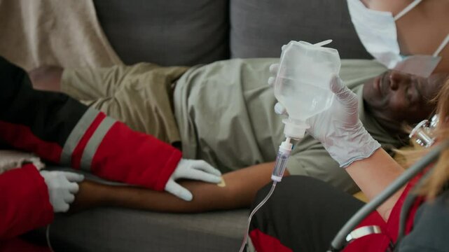 Female paramedic in protective mask and gloves holding IV bag with tube while her colleague attaching catheter to arm of Black male patient lying on couch at home during intravenous therapy