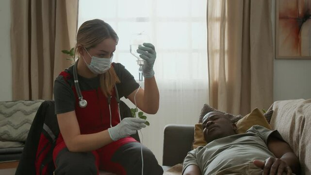 Caucasian female paramedic in sterile gloves and mask holding bag and talking to Black patient lying on couch while administering IV drip to him at home