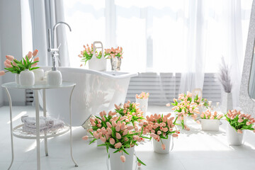 A stylish bathroom features a freestanding bathtub surrounded by pastel flowers in vases. Sunlight streams through sheer curtains, enhancing the serene atmosphere.