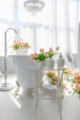 A stylish bathroom features a freestanding bathtub surrounded by pastel flowers in vases. Sunlight streams through sheer curtains, enhancing the serene atmosphere.