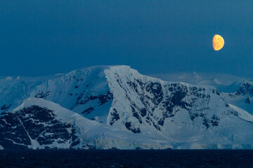 View of the nearly full moon rising over snow-covered mountains on the Antarctic Peninsula, Antarctica