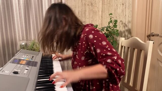 A beautiful young woman in a stylish dress and glasses plays the digital piano while practicing at home. A serene moment of creativity and dedication indoors. Hobby and leisure time