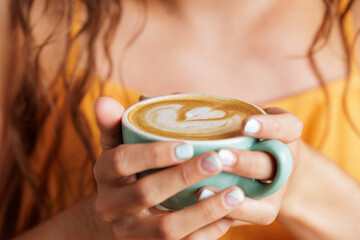 Woman's hands holding a blue cup of specialty coffee with a heart drawn on it. Woman at break time from work or student relaxing after exams.