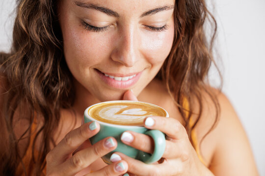 Portrait of a young woman as she smiles and enjoys the aroma of the specialty coffee she has in her hands. Coffee with a drawing of a heart. Concept of tranquility and calm, tasting a good coffee.