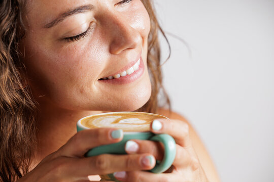 Portrait of a young woman as she smiles and enjoys the aroma of the specialty coffee she has in her hands. Coffee with a drawing of a heart. Concept of tranquility and calm, tasting a good coffee.
