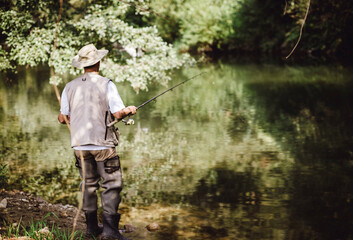 A fisherman standing by a calm river, wearing a fishing vest and hat, holding a fishing rod...