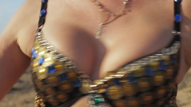 Belly dancer wearing gold and blue bra shakes her body against sunny, blurred desert backdrop, showcasing beauty and elegance of traditional dance and cultural expression