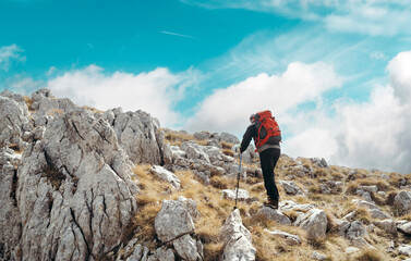 A hiker climbing a rocky mountain trail under a bright blue sky, carrying a red backpack and using trekking poles for support