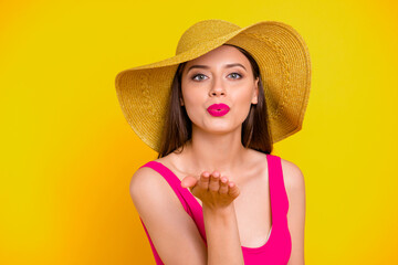 Close up portrait of girl in a brimmed hat sends an air kiss from her palm looking straight into the camera isolated on yellow background