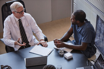 High angle shot of African American male doctor discussing with senior head physician anamnesis of patients disease sitting at meeting table, while planning treatment at medical office
